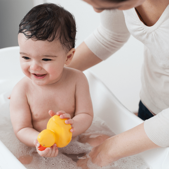 Baby in bath tub with yellow rubber duck and woman bathing him